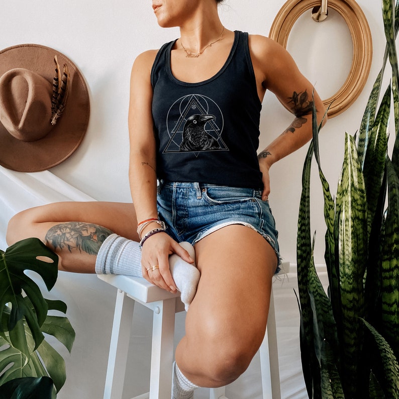 Op de afbeelding: Een zwart tanktop met een wit geometrisch ontwerp met een raaf. Het tanktop wordt gedragen door een persoon met een tatoeage op hun arm en een witte sok op hun voet.