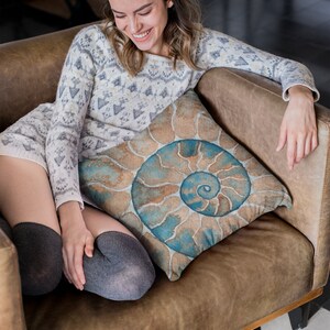 May include: A woman sits on a brown leather couch with a throw pillow featuring a blue and brown spiral pattern. She is wearing a white and gray sweater and gray tights.