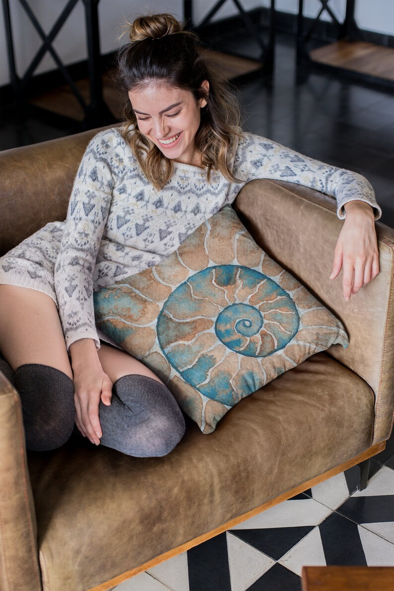 May include: A woman sits on a brown leather couch with a throw pillow featuring a blue and brown spiral pattern. She is wearing a white and gray sweater and gray tights.