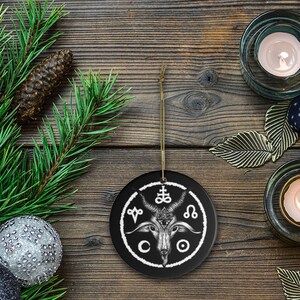May include: Black and white ceramic Christmas ornament featuring a goat head with horns and a pentagram above it. The ornament is surrounded by a circle with symbols and the letters "CO" at the bottom.