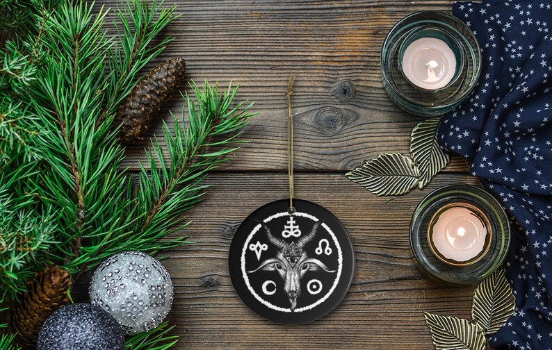 May include: Black and white ceramic Christmas ornament featuring a goat head with horns and a pentagram above it. The ornament is surrounded by a circle with symbols and the letters "CO" at the bottom.