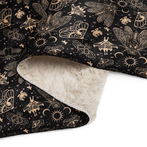 May include: Black blanket with a repeating pattern of insects, moons, and suns in tan and pink. The blanket has a soft, cream-coloured sherpa lining. The design features moths, cicadas, and other stylised insects.