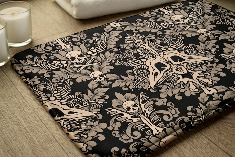 May include: A black bath mat with a beige damask pattern featuring skulls and crossbones, skeletons, and floral designs.