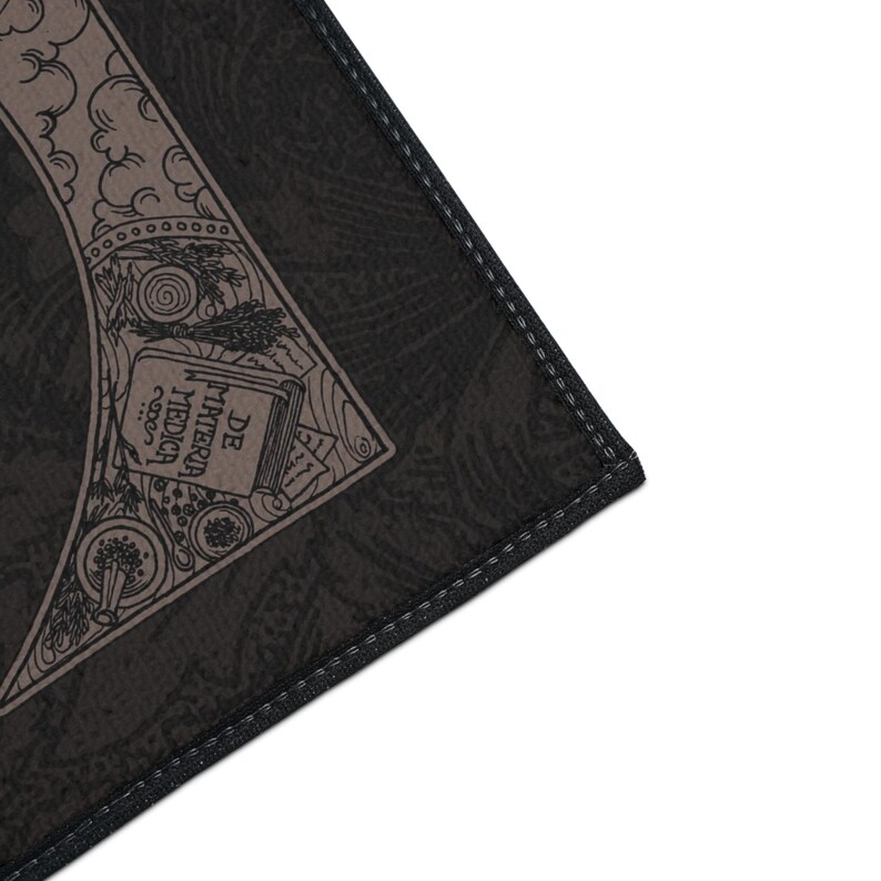 May include: Close-up of a black and brown patterned fabric with a stitched edge. The fabric features a vintage illustration of a book titled "DE MATERIA MEDICA".