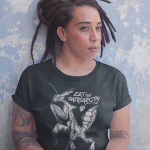 Może przedstawiać: Czarny t-shirt z białym nadrukiem przedstawiającym modliszkę i tekstem "Eat the Patriarchy" z symbolem żeńskim.