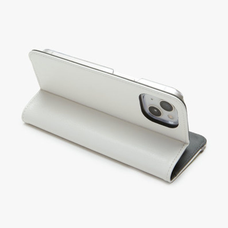 May include: White leather phone case with a black flap. The case is closed and the camera lens is visible.