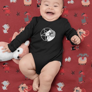 May include: A baby wearing a black long-sleeve onesie with a white pentagram and goat head graphic. The baby is lying on a red blanket with a repeating pattern of white goats and stars.