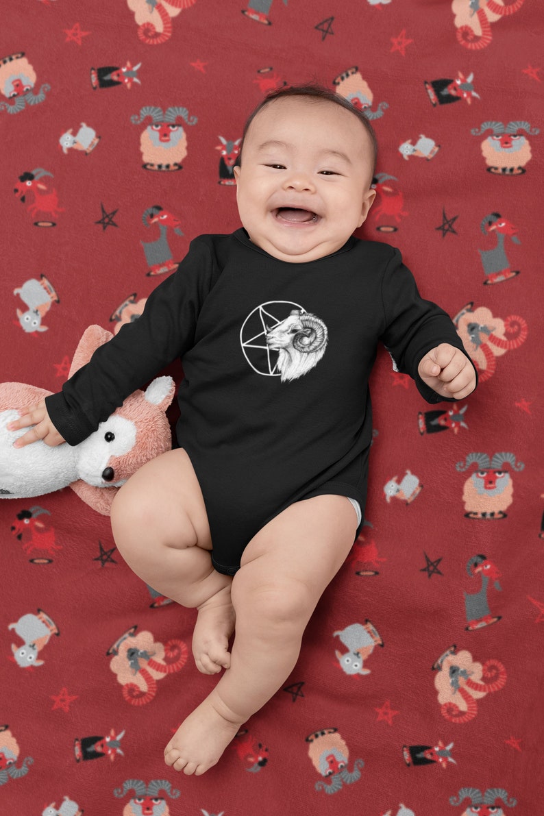 May include: A baby wearing a black long-sleeve onesie with a white pentagram and goat head graphic. The baby is lying on a red blanket with a repeating pattern of white goats and stars.