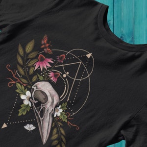May include: A black t-shirt with a graphic design featuring a bird skull surrounded by pink and white flowers, butterflies, and geometric shapes.