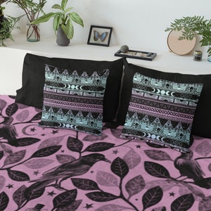 May include: A pink and black patterned bedding set with a design of ravens and leaves. The set includes two throw pillows with a black background and a geometric pattern in shades of blue, pink, and white.