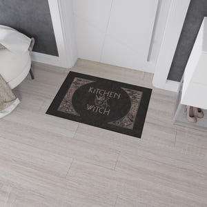 May include: Black doormat with the text "Kitchen Witch" in a decorative font. The text is surrounded by a circle with a cauldron and other witchy symbols.