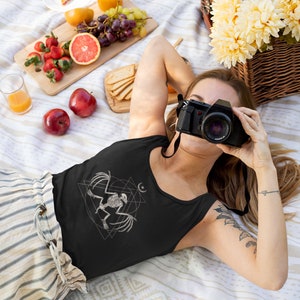 May include: A woman wearing a black tank top with a white graphic of a bird skeleton inside a geometric design. She is lying on a white and brown blanket with a camera in front of her face.