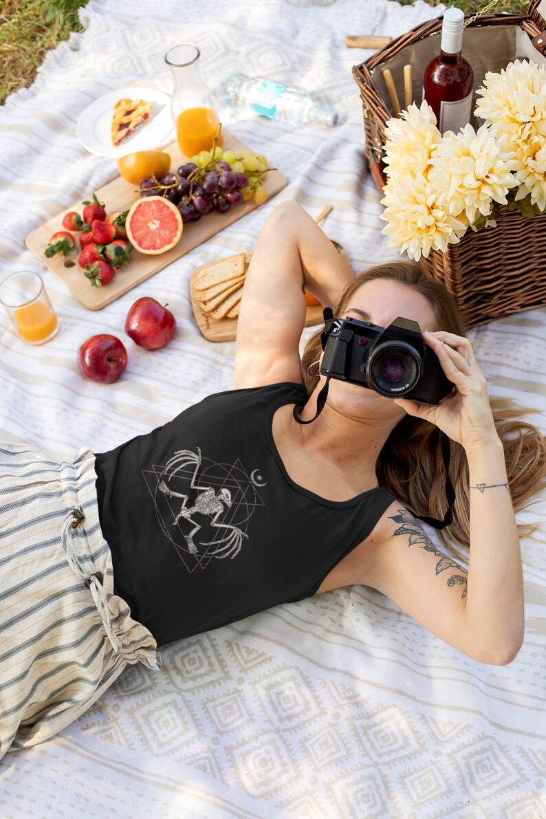 May include: A woman wearing a black tank top with a white graphic of a bird skeleton inside a geometric design. She is lying on a white and brown blanket with a camera in front of her face.