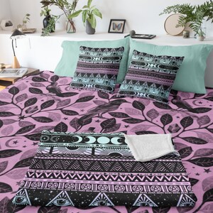 May include: A pink and black patterned throw blanket with a geometric design featuring moons, stars, and eyes. The blanket is on a bed with matching pillowcases.