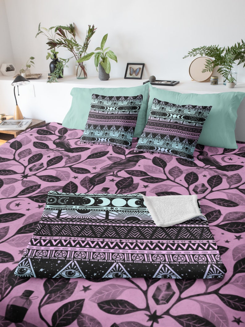 May include: A pink and black patterned throw blanket with a geometric design featuring moons, stars, and eyes. The blanket is on a bed with matching pillowcases.