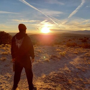May include: A person wearing a black hoodie with a white graphic on the back, standing on a sandy hill, looking at a sunset. The sky is filled with clouds and contrails, with the sun low on the horizon. The landscape is a desert scene.