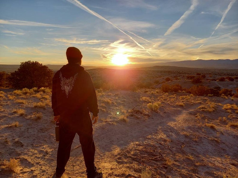 May include: A person wearing a black hoodie with a white graphic on the back, standing on a sandy hill, looking at a sunset. The sky is filled with clouds and contrails, with the sun low on the horizon. The landscape is a desert scene.