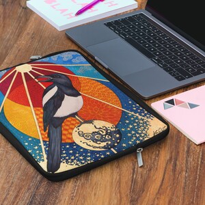 May include: A colorful laptop sleeve with a black magpie perched on a sphere with a sun and clouds in the background.