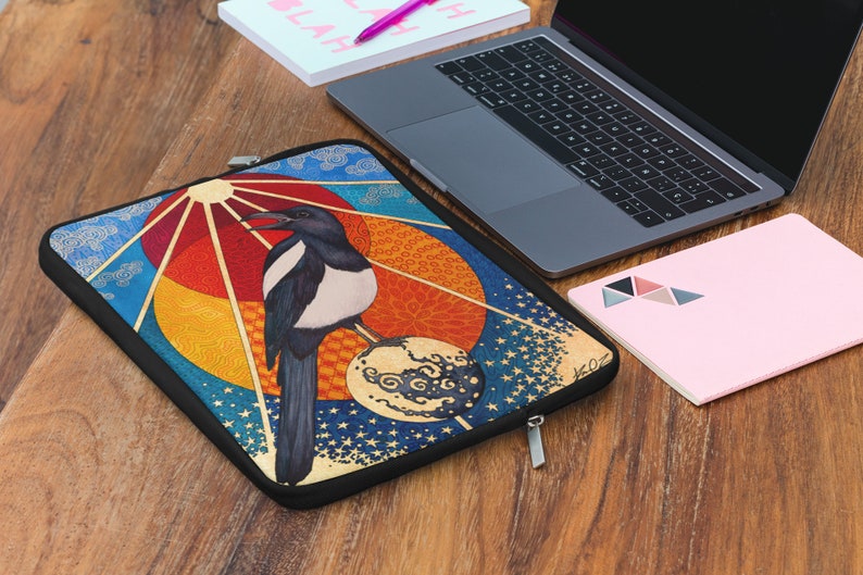 May include: A colorful laptop sleeve with a black magpie perched on a sphere with a sun and clouds in the background.