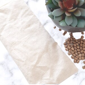 May include: A white cotton eye pillow filled with small brown seeds. The pillow is lying on a white marble surface.