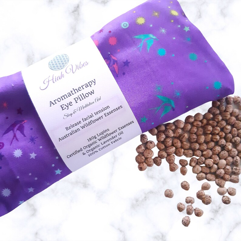 May include: Purple aromatherapy eye pillow with a floral print and the text "Hiah Vibes Aromatherapy Eye Pillow Step & Meditation Aid Release facial tension Australian Wildflower Essenses 180g Lupins Certified Organic Wildflower Essenses & Organic Lavender Oil 100% Cotton Fabric". The pillow is filled with brown lupin seeds.