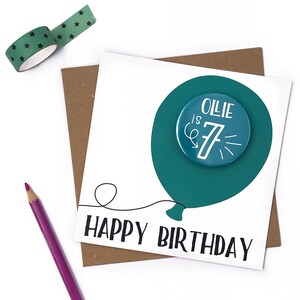 May include: A white card with a teal balloon with a button that says "Ollie is 7" on it. The card says "Happy Birthday" in black text.