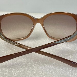 K&ouml;nnte beinhalten: Braune ovale Sonnenbrille mit Farbverlaufgl&auml;sern. Die Fassungen sind hellbraun, mit glitzernden Verzierungen an den B&uuml;geln. Die B&uuml;gel sind dunkelbraun und &uuml;berkreuzt.
