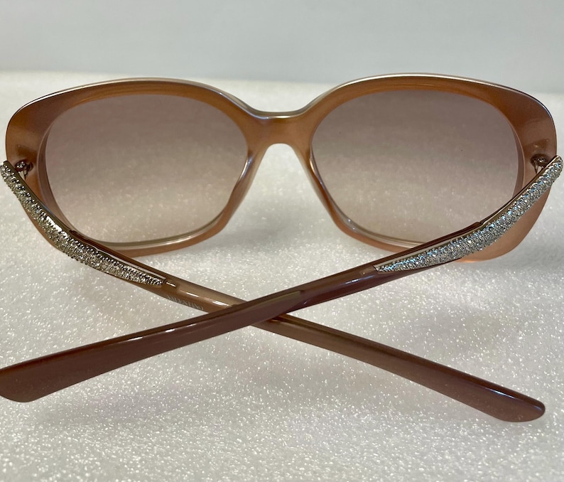 K&ouml;nnte beinhalten: Braune ovale Sonnenbrille mit Farbverlaufgl&auml;sern. Die Fassungen sind hellbraun, mit glitzernden Verzierungen an den B&uuml;geln. Die B&uuml;gel sind dunkelbraun und &uuml;berkreuzt.
