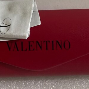 K&ouml;nnte beinhalten: Ein rotes Valentino-Brillenetui mit einem passenden wei&szlig;en Reinigungstuch. Das Etui ist rechteckig mit gew&ouml;lbtem Deckel und der Markenname ist schwarz aufgedruckt. Das Tuch hat ein aufgedrucktes Logo.