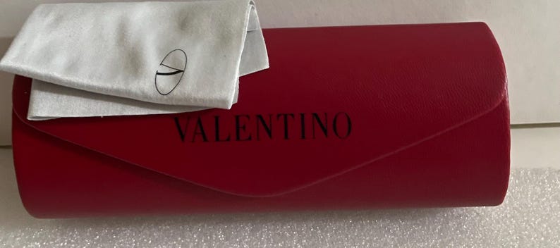 K&ouml;nnte beinhalten: Ein rotes Valentino-Brillenetui mit einem passenden wei&szlig;en Reinigungstuch. Das Etui ist rechteckig mit gew&ouml;lbtem Deckel und der Markenname ist schwarz aufgedruckt. Das Tuch hat ein aufgedrucktes Logo.