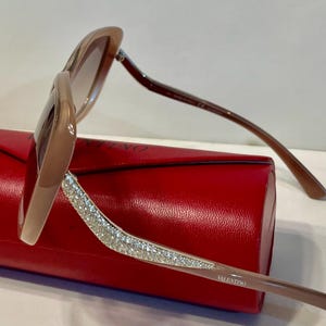 K&ouml;nnte beinhalten: Braune Sonnenbrille mit kristallbesetztem B&uuml;gel, auf einem roten Etui liegend. Die Sonnenbrille hat einen hellbraunen Rahmen und Verlaufsgl&auml;ser. Der Markenname "VALENTINO" ist auf dem B&uuml;gel zu sehen.
