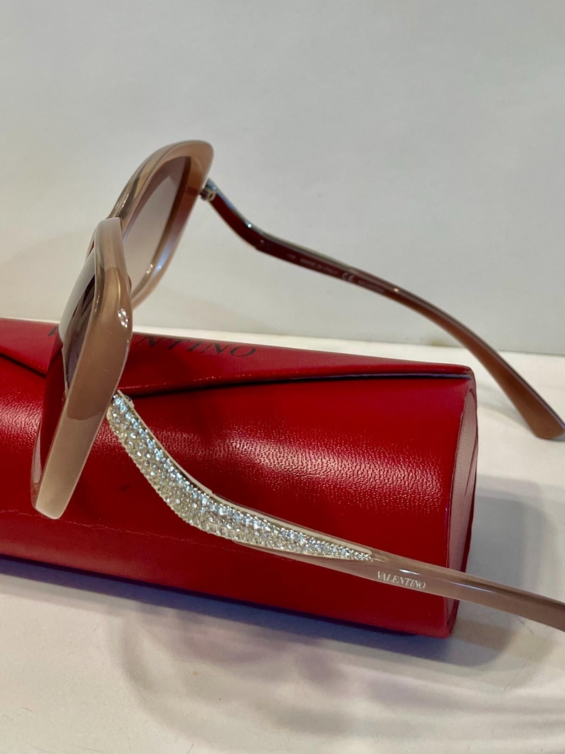 K&ouml;nnte beinhalten: Braune Sonnenbrille mit kristallbesetztem B&uuml;gel, auf einem roten Etui liegend. Die Sonnenbrille hat einen hellbraunen Rahmen und Verlaufsgl&auml;ser. Der Markenname "VALENTINO" ist auf dem B&uuml;gel zu sehen.