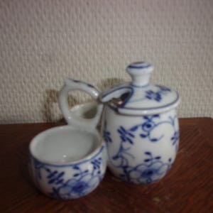 Könnte beinhalten: Ein weißes Keramik-Salz- und Pfefferset mit blauen Blumenmustern. Das Set enthält eine kleine offene Schüssel mit Griff und ein Gefäß mit Deckel und Löffelablage. Die Teile stehen auf einer Holzoberfläche.