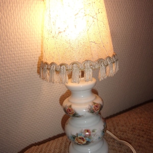 Può includere: Una lampada da tavolo vintage con base in ceramica bianca decorata con motivi floreali rosa, gialli e blu. La lampada presenta un paralume strutturato con finiture e nappe decorative. La lampada è collegata e accesa.