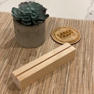 May include: A wooden sign holder with a slot for displaying a card or sign. The holder is made of natural wood and has a rustic look. It is sitting on a woven mat with a small potted succulent plant and a round wooden coaster with the text "Laser Craft Fabrications" in the center.