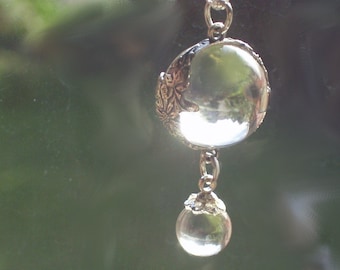 Puede incluir: Una cadena de plata con dos orbes de vidrio transparente colgando de ella. El orbe más grande tiene un diseño floral decorativo.