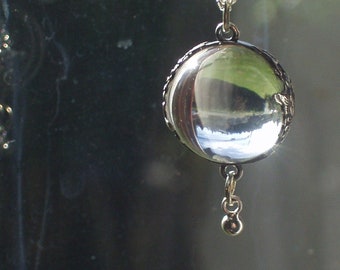 May include: A silver-toned pendant necklace featuring a round, clear orb that reflects a landscape scene. The orb is encased in a decorative metal frame and hangs from a delicate chain. A small, dangling bead is suspended below the orb.