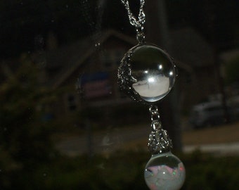 Puede incluir: Un collar de cadena de plata con un gran colgante de cristal transparente y un colgante de ópalo más pequeño. El orbe más grande refleja el entorno circundante.