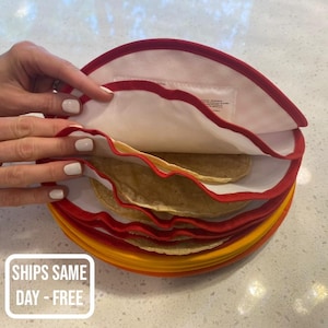 May include: A stack of round, flat, light brown tortillas are stored in a red and yellow tortilla keeper. The keeper has a white fabric lining with a red trim.