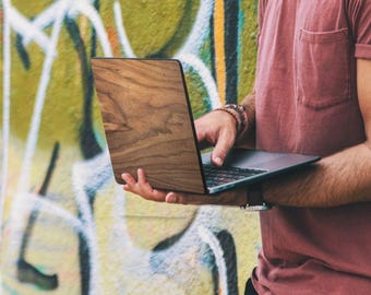 Puede incluir: Una persona sostiene un portátil con carcasa de madera y pantalla oscura. Viste una camiseta roja y pantalones negros, de pie frente a una pared cubierta de grafitis. Se aprecia la veta de la madera del portátil.