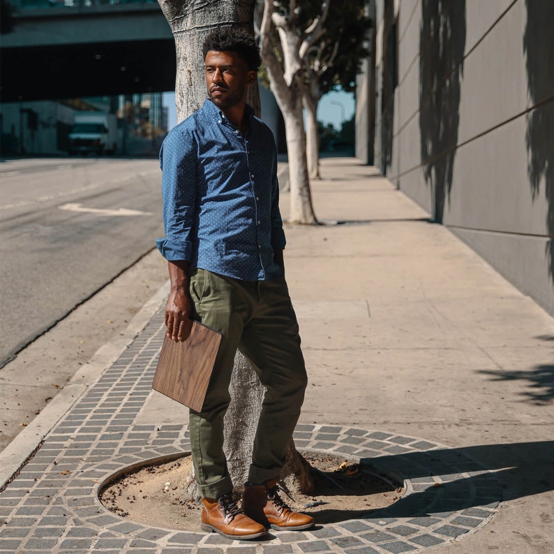 Puede incluir: Una persona con una camisa azul con lunares blancos, pantalones verde oliva y botas de cuero marrones. Sostiene un objeto cuadrado de madera. La escena es al aire libre, junto a un &aacute;rbol y una acera.