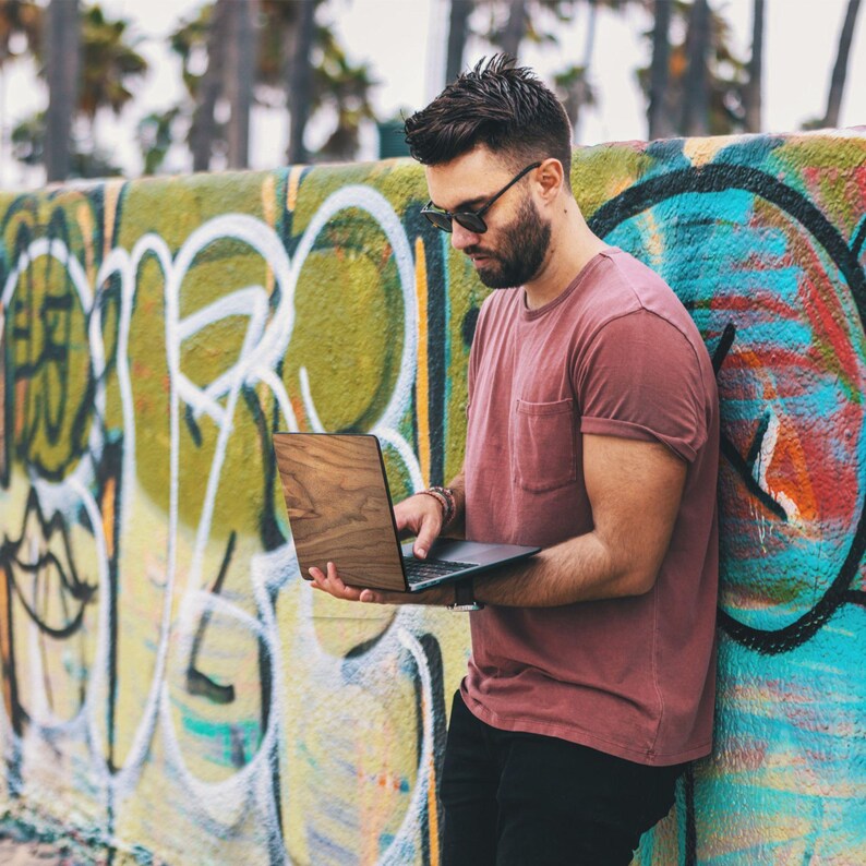 Puede incluir: Una persona sostiene un port&aacute;til con carcasa de madera y pantalla oscura. Viste una camiseta roja y pantalones negros, de pie frente a una pared cubierta de grafitis. Se aprecia la veta de la madera del port&aacute;til.