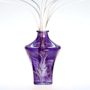 Può includere: Un vaso di vetro viola con la parte superiore svasata e disegni floreali incisi. Il vaso contiene canne bianche, creando un'esposizione decorativa. Il vaso è di colore viola scuro e si erge su uno sfondo bianco.