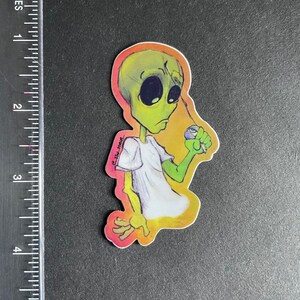 Peut inclure: Un autocollant représentant un extraterrestre vert avec une expression triste, tenant un yo-yo bleu et blanc. L'extraterrestre porte un t-shirt blanc et est sur un fond orange, jaune et rose.