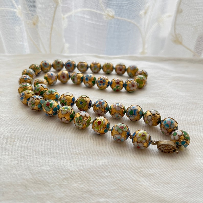 May include: A gold necklace with round beads decorated with colorful enamel floral patterns. The beads are strung on a gold chain and have a silver clasp.