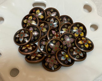 May include: A collection of 15 small, round, black enamel buttons with colorful floral designs. The buttons are arranged in a circle on a white surface.