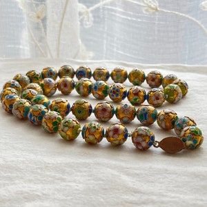 May include: A gold necklace with round beads decorated with colorful enamel flowers. The beads are connected with a thin gold chain.