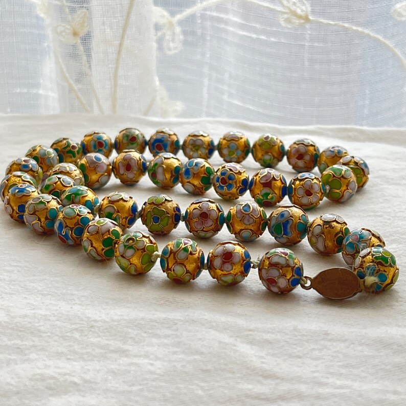 May include: A gold necklace with round beads decorated with colorful enamel flowers. The beads are connected with a thin gold chain.