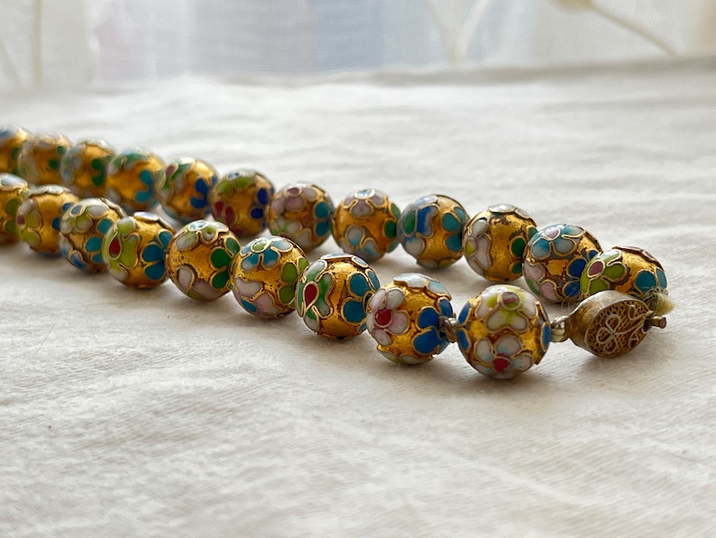 May include: A gold necklace with round beads decorated with colorful enamel floral patterns. The beads are connected with a silver clasp.