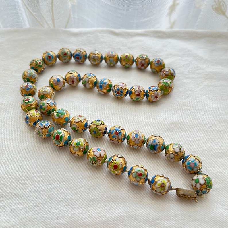 May include: A gold necklace with round beads decorated with colorful enamel floral patterns. The beads are strung on a gold chain and have a clasp closure.